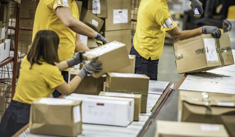 Group of postal workers handling and sorting packages at a conveyor belt in a warehouse.