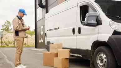 Young man in uniform using his mobile phone while standing near the van and packages outdoors near the warehouse for last-mile delivery