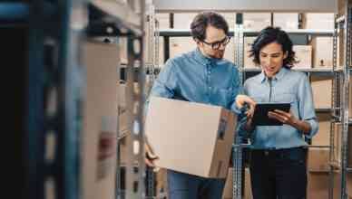 Female Inventory Manager Shows Delivery Management Software to a Worker Holding Cardboard Box, They Talk and Do Work. In the Background Stock of Parcels with Products Ready for Shipment.