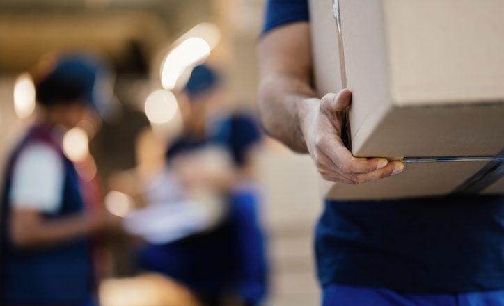 Close-up of a worker carrying cardboard box while making a delivery. There are people in the background.