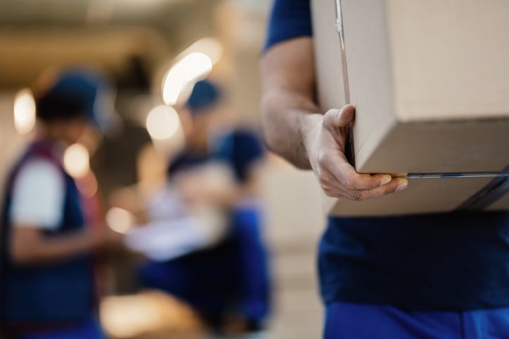 Close-up of a worker carrying cardboard box while making a delivery. There are people in the background.