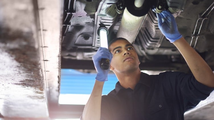 A young mechanic is working under a car in a garage repair shop. He is wearing blue overalls. He is looking up with an inspection lamp to check the possible damage that has been sustained to the exhaust.