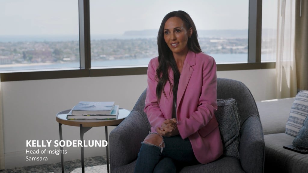 Kelly Soderlund, Head of Insights at Samsara, sits in an office chair wearing a pink blazer and jeans, speaking with hands clasped, with cityscape visible through large windows behind her.