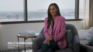 Kelly Soderlund, Head of Insights at Samsara, sits in an office chair wearing a pink blazer and jeans, speaking with hands clasped, with cityscape visible through large windows behind her.