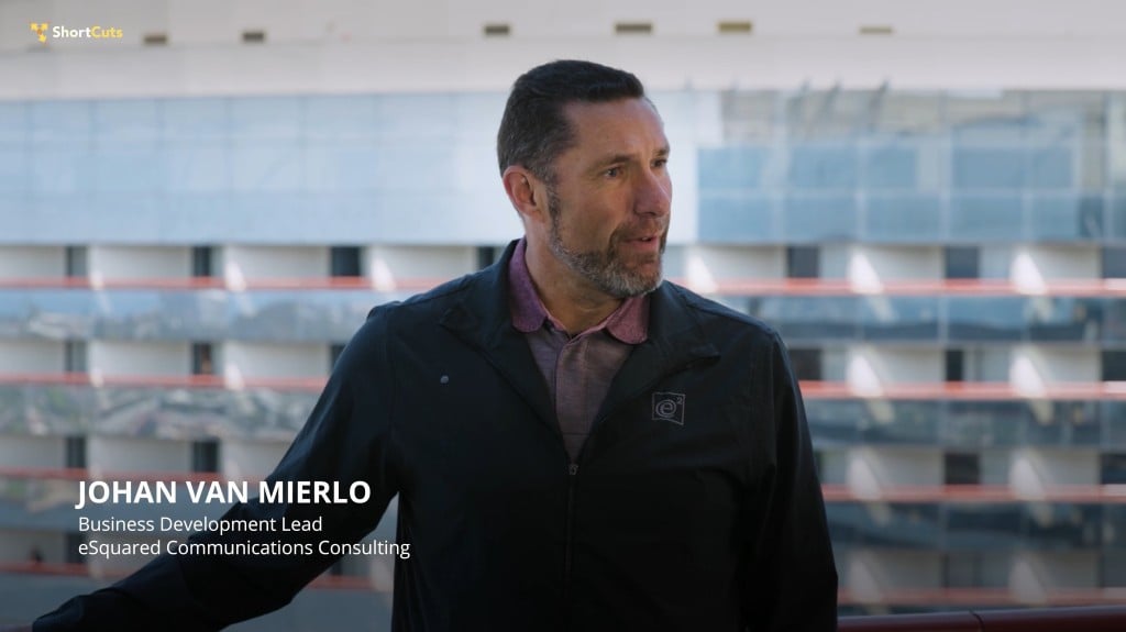 Johan Van Mierlo, Business Development Lead at eSquared Communications Consulting, speaks outdoors in a dark jacket with the eSquared logo, captioned on screen with a blurred building and āShortCutsā logo.