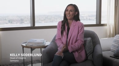 Kelly Soderlund, Head of Insights at Samsara, sits in an office chair wearing a pink blazer and jeans, speaking with hands clasped, with cityscape visible through large windows behind her.