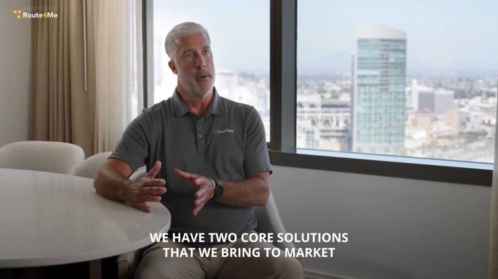 Tom Robertson, wearing a gray PrePass polo, speaks while seated at a round table in a high-rise office with city buildings visible through the window. On-screen text reads: "We have two core solutions that we bring to market."