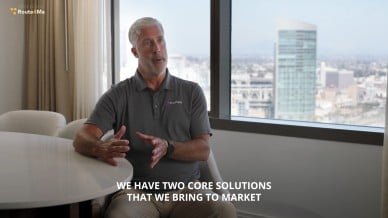 Tom Robertson, wearing a gray PrePass polo, speaks while seated at a round table in a high-rise office with city buildings visible through the window. On-screen text reads: "We have two core solutions that we bring to market."