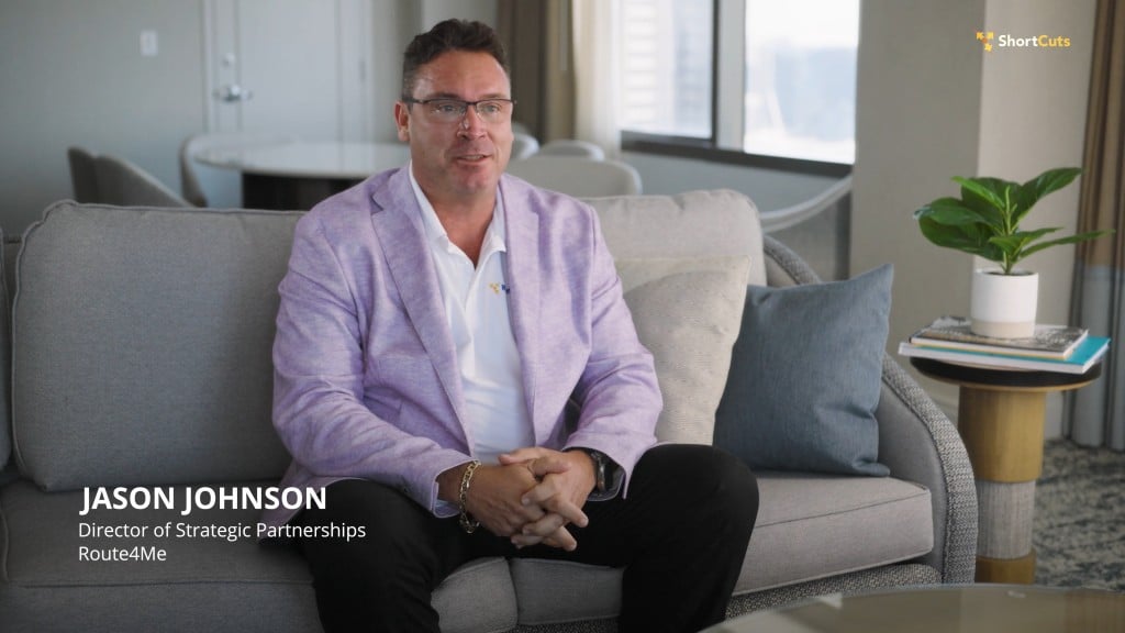 Jason Johnson, Director of Strategic Partnerships at Route4Me, sits on a gray couch in a light purple blazer, white shirt, and black pants, speaking during a Route4Me ShortCuts interview.