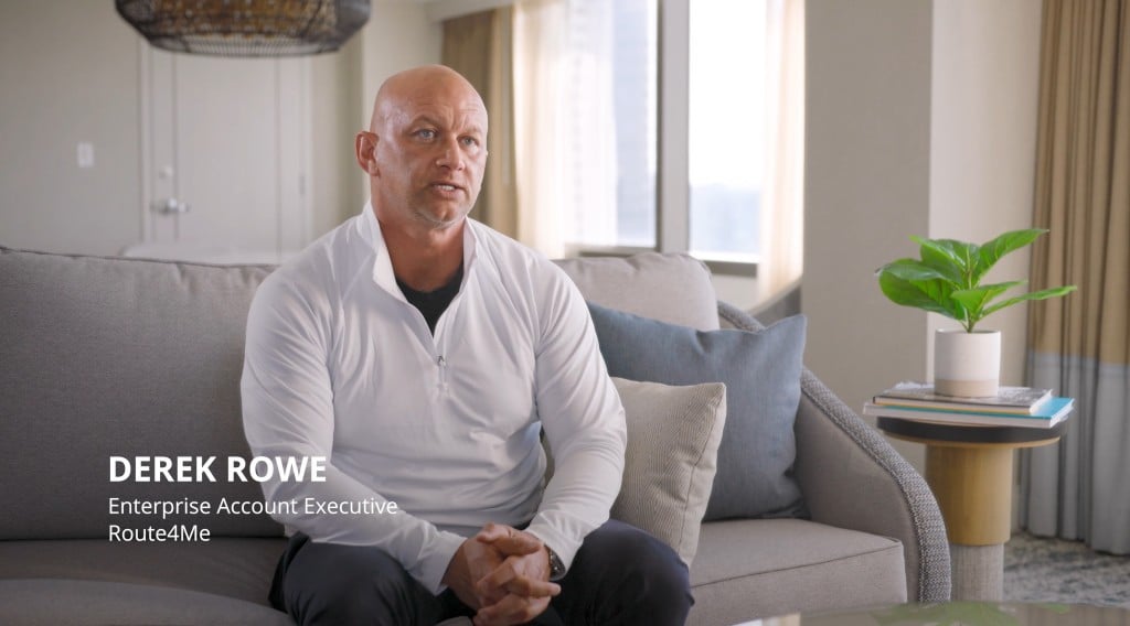 Derek Rowe, Enterprise Account Executive at Route4Me, sits on a gray sofa in a bright office setting, wearing a white long sleeve shirt, with hands clasped. A small plant and stacked books rest on a side table next to him.