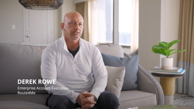 Derek Rowe, Enterprise Account Executive at Route4Me, sits on a gray sofa in a bright office setting, wearing a white long sleeve shirt, with hands clasped. A small plant and stacked books rest on a side table next to him.