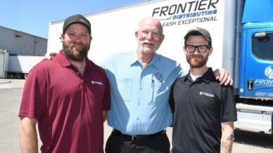 Three men smile arm-in-arm before a white Frontier Distributing truck and blue Frontier semi cab on a sunny day.