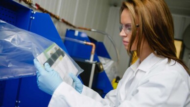 SoBran technician in lab coat, safety glasses, and gloves inspecting a sealed clear package at a screening station
