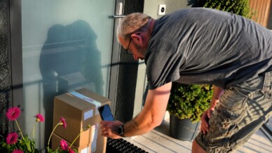 A man bends down to photograph or scan a cardboard package left at a glass front door with his phone, pink flowers nearby in the sunlight.