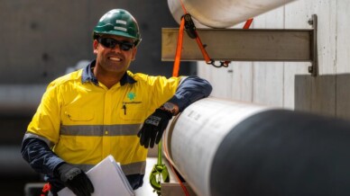 Smiling field worker in yellow hi-vis and green hard hat leans against a large industrial pipe at a utility site.