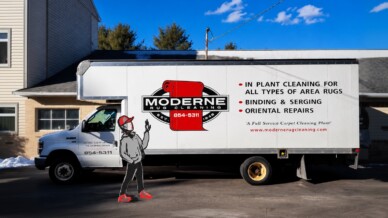 A white Moderne Rug Cleaning box truck parked outside a building, with an illustrated cartoon figure in a red cap waving beside it.